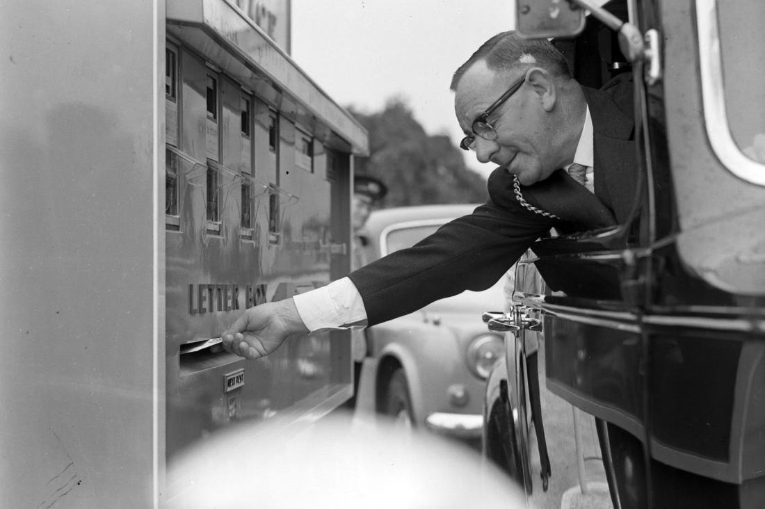 Black and white ohoto of man posting a letter from his car window 