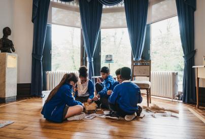 Four children weraring school uniforms sitting inside a well-lit space inside Wardown House, Museum & Gallery
