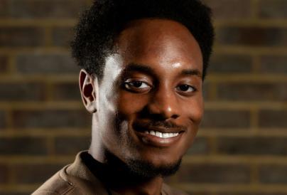 Kevon Anokwawe - young black male, sporting medium curled hair with a relaxed smile on his face, wearing a brown shirt in front of a blurred brick wall 