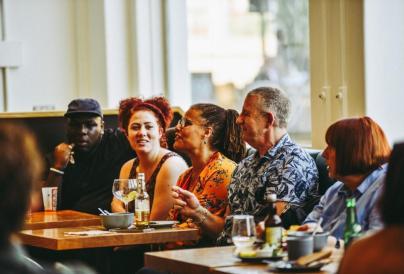 A group of adults sit together around tables in a bright café or bar, chatting and smiling during a social event.
