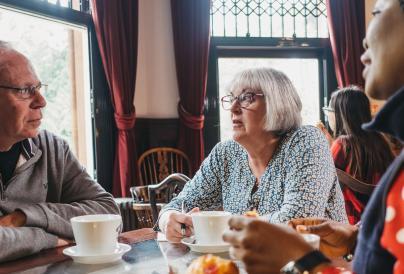 A group of people sit around a table in a bright café, chatting over cups of coffee in a relaxed, welcoming setting.