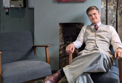 A young man dressed in a smart grey three-piece suit and burgundy tie sits confidently in an armchair inside a vintage-style interior.