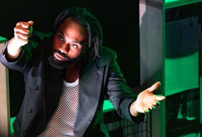 Man wearing black blazer and white mesh top in front of a background of boxes lit up in green