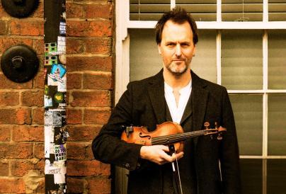 A man stands in front of a brick wall, holding a violin with a focused expression.